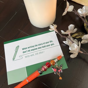May include: A motivational quote card with a green and white background. The quote reads "When writing the story of your life, don't let anyone else hold your pen. Write well, Edit often!" An orange pen with colorful beads and a butterfly charm is on top of the card.