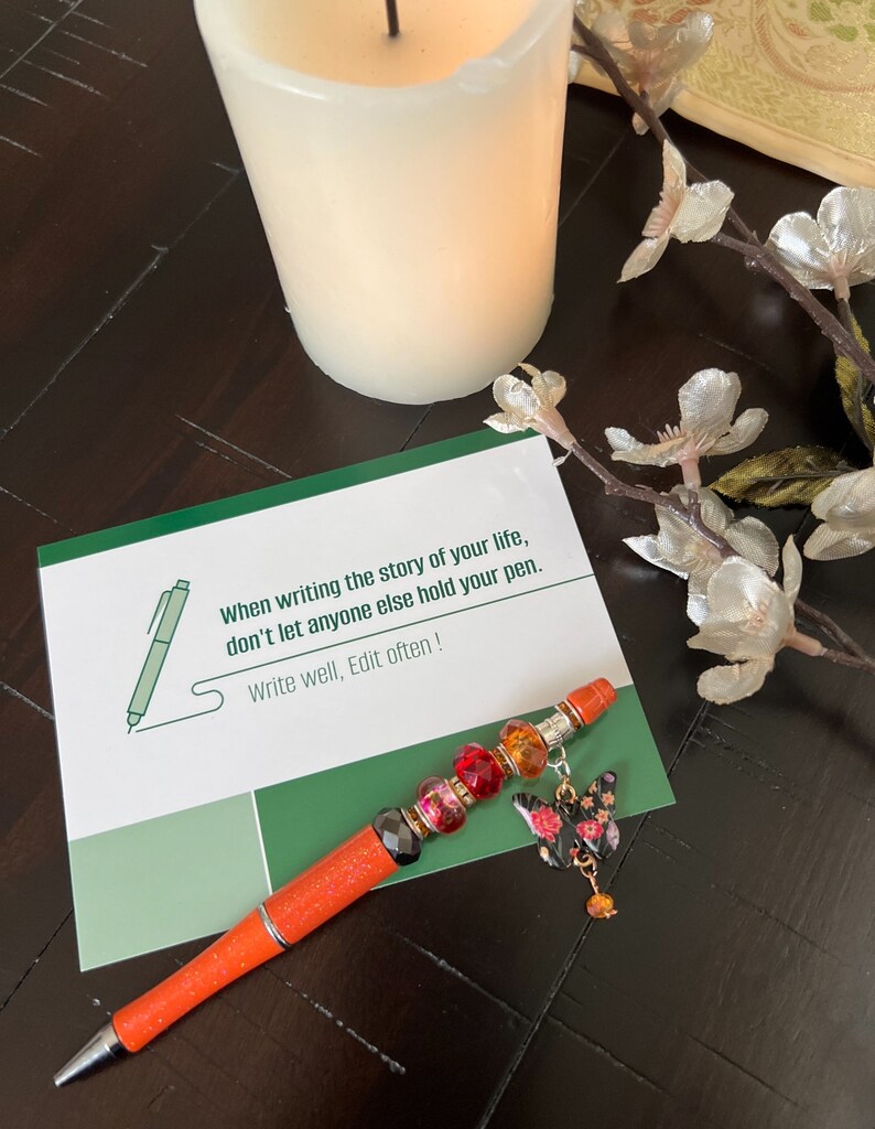 May include: A motivational quote card with a green and white background. The quote reads "When writing the story of your life, don't let anyone else hold your pen. Write well, Edit often!" An orange pen with colorful beads and a butterfly charm is on top of the card.