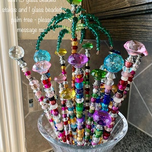May include: A glass vase filled with 15 colourful glass beaded stakes and a single glass beaded palm tree. The stakes are topped with various coloured glass beads and the palm tree is topped with a green glass bead.