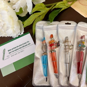 May include: Five colorful pens with decorative embellishments are displayed in individual clear plastic bags. Each pen has a different design featuring beads, charms, and other decorative elements. The pens are packaged for sale.