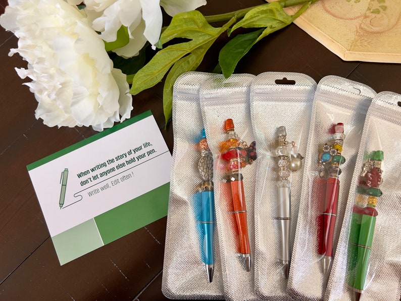 May include: Five colorful pens with decorative embellishments are displayed in individual clear plastic bags. Each pen has a different design featuring beads, charms, and other decorative elements. The pens are packaged for sale.