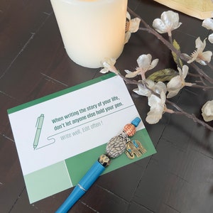 May include: A blue pen with a decorative elephant charm and colorful beads. The pen is lying on a card with the text "When writing the story of your life, don't let anyone else hold your pen. Write well, Edit often!"