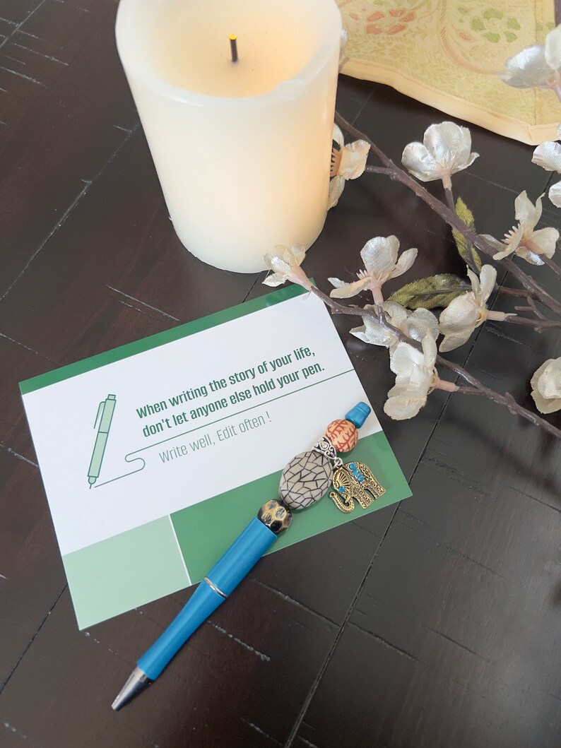 May include: A blue pen with a decorative elephant charm and colorful beads. The pen is lying on a card with the text "When writing the story of your life, don't let anyone else hold your pen. Write well, Edit often!"