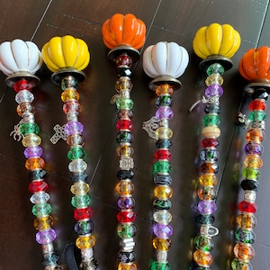 May include: Six decorative pumpkin-shaped garden stakes with colorful beads and charms. The pumpkins are white, yellow, and orange. The beads are red, green, purple, and clear. The charms include a spider, a cross, and a small metal charm.