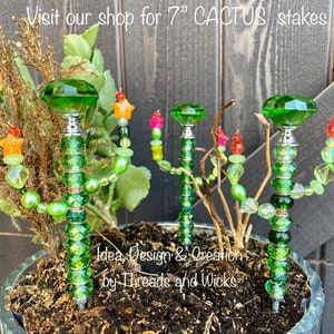K&ouml;nnte beinhalten: Vier 17,8 cm lange gr&uuml;ne Glas-Kaktus-Stiche mit gr&uuml;nen, gelben und roten Perlen. Die Stiche befinden sich in einem Topf mit brauner Erde. Der Text "Idea, Design & Creation by Threads and Wicks" ist im Bild sichtbar.