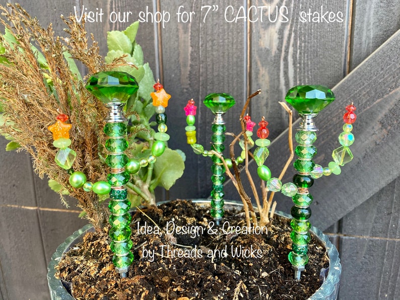 K&ouml;nnte beinhalten: Vier 17,8 cm lange gr&uuml;ne Glas-Kaktus-Stiche mit gr&uuml;nen, gelben und roten Perlen. Die Stiche befinden sich in einem Topf mit brauner Erde. Der Text "Idea, Design & Creation by Threads and Wicks" ist im Bild sichtbar.