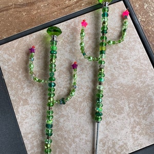 May include: Two green beaded cactus garden stakes with pink and purple flowers. The stakes are made of metal and have a green glass bead at the top.