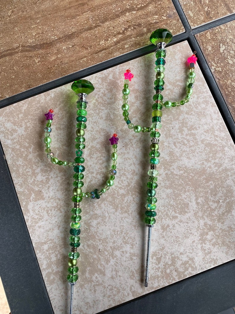 May include: Two green beaded cactus garden stakes with pink and purple flowers. The stakes are made of metal and have a green glass bead at the top.