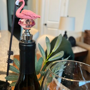 Pode incluir: Tampa de garrafa de flamingo rosa com base de cristal. A tampa est&aacute; no topo de uma garrafa de vinho.