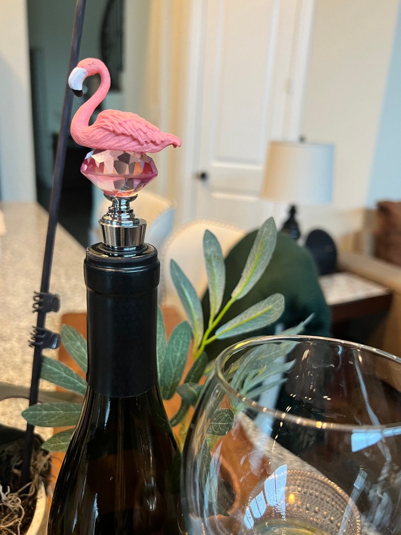 Pode incluir: Tampa de garrafa de flamingo rosa com base de cristal. A tampa est&aacute; no topo de uma garrafa de vinho.