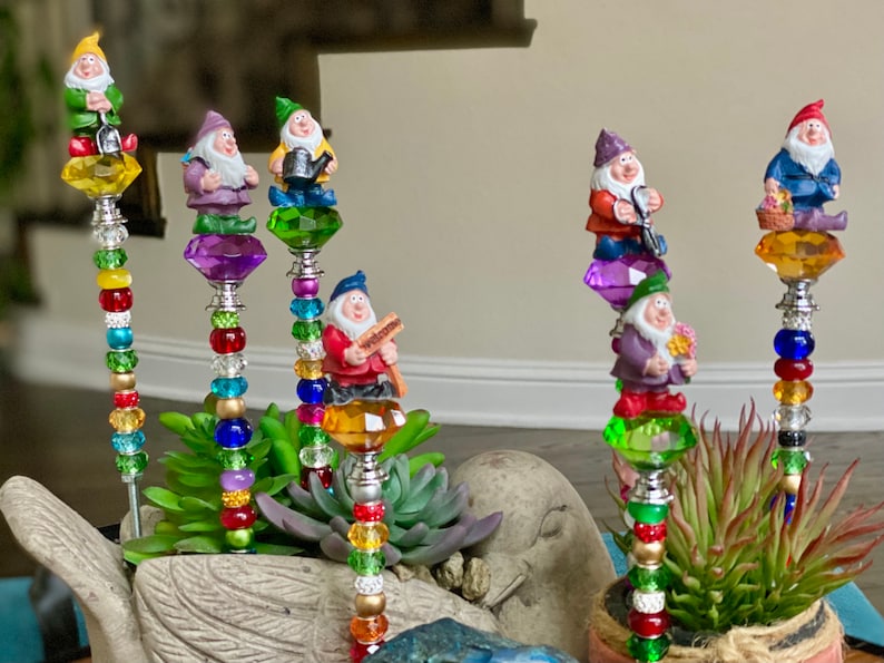K&ouml;nnte beinhalten: Sechs bunte Gartenst&auml;be mit Gnomfiguren oben drauf. Jeder Stab ist mit bunten Perlen und Kristallen verziert. Die Gnome tragen verschiedene Outfits und halten verschiedene Gartenwerkzeuge in der Hand.
