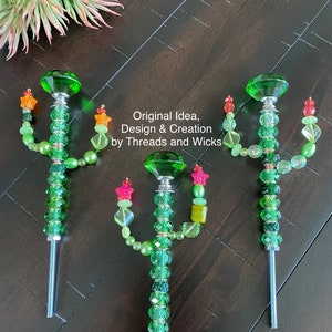 May include: Three green glass cactus-shaped garden stakes with colorful beads and star-shaped accents. The stakes are made of metal and have a screw-on top. The text "Original Idea, Design & Creation by Threads and Wicks" is visible in the image.