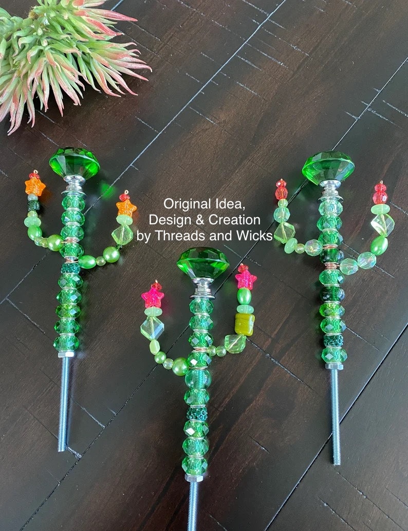 May include: Three green glass cactus-shaped garden stakes with colorful beads and star-shaped accents. The stakes are made of metal and have a screw-on top. The text "Original Idea, Design & Creation by Threads and Wicks" is visible in the image.