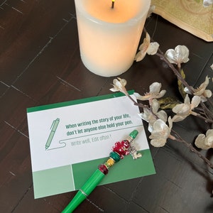 May include: A green pen with red, gold, and silver beads and a charm. The pen is lying on a green card with the text "When writing the story of your life, don't let anyone else hold your pen. Write well, Edit often!"