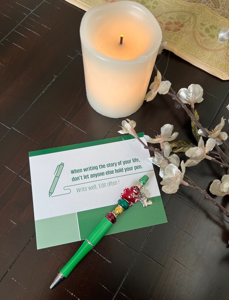 May include: A green pen with red, gold, and silver beads and a charm. The pen is lying on a green card with the text "When writing the story of your life, don't let anyone else hold your pen. Write well, Edit often!"