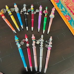 May include: A collection of 15 colorful pens with various charms and beads. The pens are arranged in two rows on a dark wooden surface. The charms include hearts, stars, flowers, and other decorative elements.