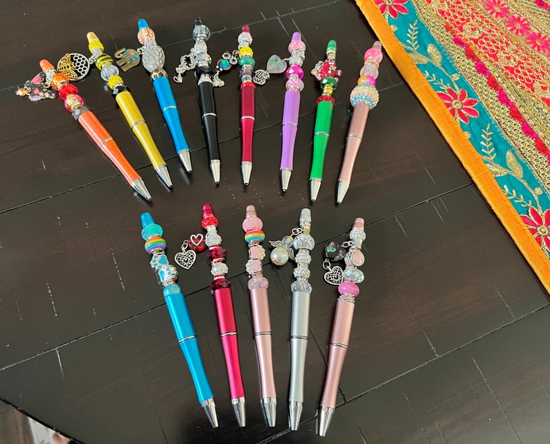 May include: A collection of 15 colorful pens with various charms and beads. The pens are arranged in two rows on a dark wooden surface. The charms include hearts, stars, flowers, and other decorative elements.