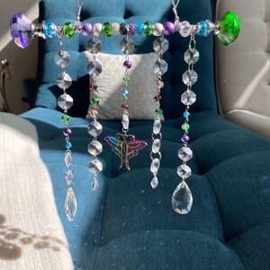 May include: A crystal suncatcher with a silver metal bar and colorful beads. The suncatcher has five strands of crystals, each with a different length. The crystals are clear and have a faceted design. The beads are green, purple, and silver. A butterfly charm hangs from the center of the bar.