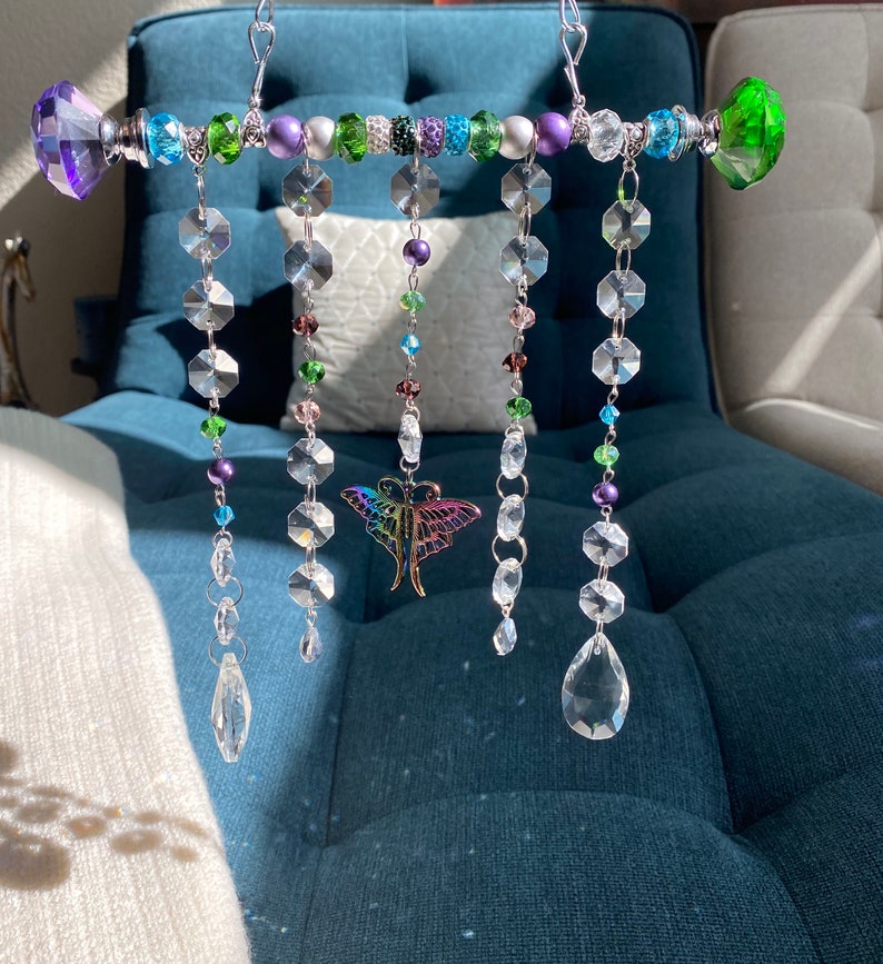 May include: A crystal suncatcher with a silver metal bar and colorful beads. The suncatcher has five strands of crystals, each with a different length. The crystals are clear and have a faceted design. The beads are green, purple, and silver. A butterfly charm hangs from the center of the bar.