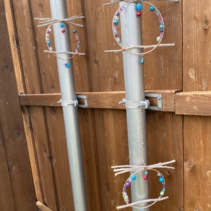 May include: Three colorful beaded wind chimes hang from metal poles attached to a wooden fence. Each chime is made of twigs and beads in shades of blue, green, pink, and red.