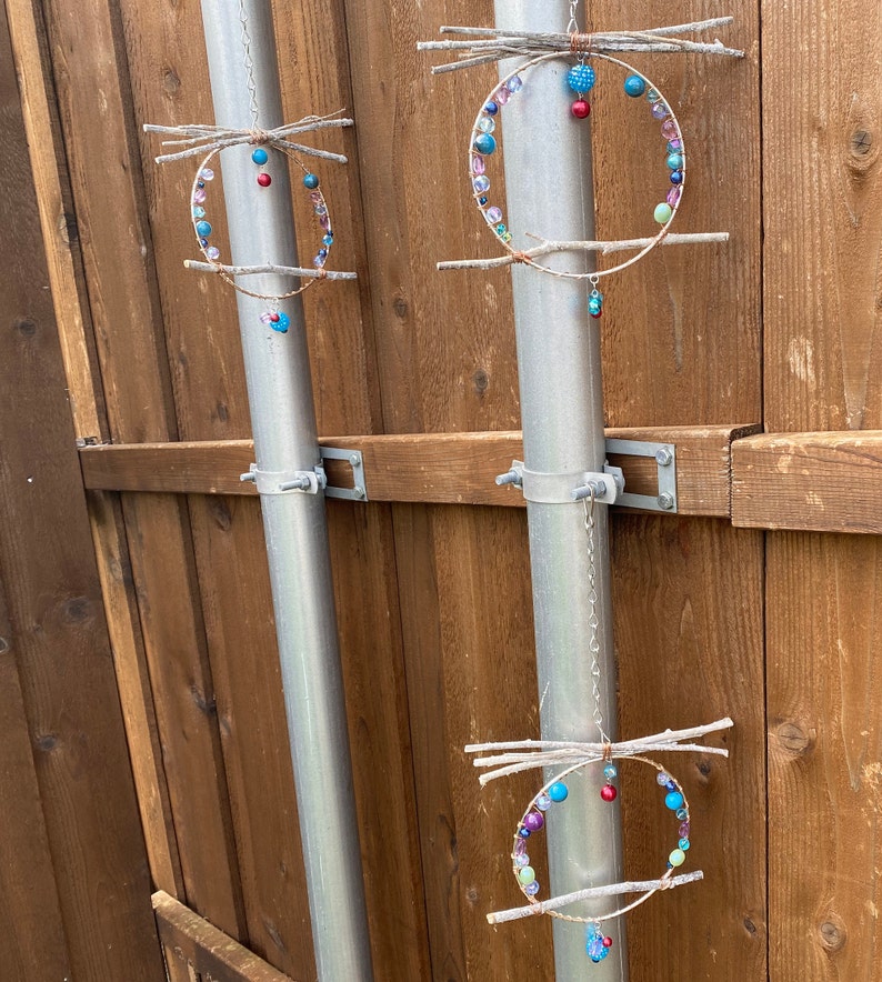 May include: Three colorful beaded wind chimes hang from metal poles attached to a wooden fence. Each chime is made of twigs and beads in shades of blue, green, pink, and red.