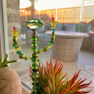 May include: A green glass cactus with a large green crystal on top. The cactus is decorated with green and yellow beads and stars. The cactus is in a pot with a succulent plant.