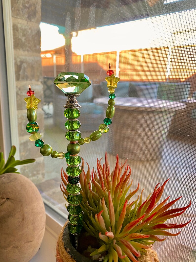 May include: A green glass cactus with a large green crystal on top. The cactus is decorated with green and yellow beads and stars. The cactus is in a pot with a succulent plant.