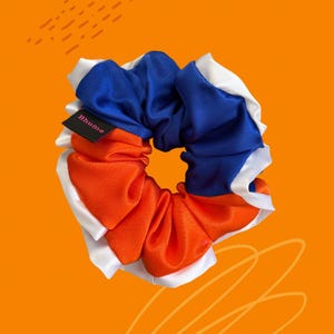 May include: A circular hair scrunchie featuring a color-blocked design of royal blue and bright orange fabric, accented with a white ruffled trim along the edges. A small black fabric tag with the pink text Bhumo is attached to the side.