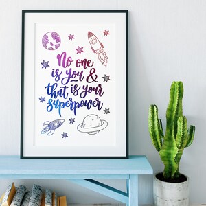 May include: A framed print featuring a motivational quote in a rainbow gradient: "No one is you & that is your superpower." The quote is surrounded by stars and rocket ship illustrations.