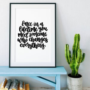 May include: Black and white framed print with the quote "Once in a lifetime you meet someone who changes everything." The print is hanging on a white wall above a light blue bench with a potted cactus.