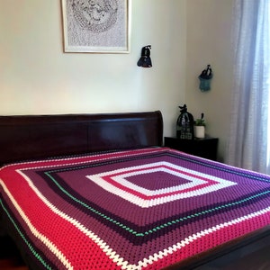 May include: A crocheted bedspread with a square pattern in shades of pink, purple, white, and green. The bedspread is on a wooden bed frame.