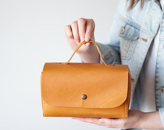 May include: A small, rectangular, tan leather handbag with a top handle and a single button closure.