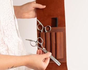 Puede incluir: Una funda de cuero marrón con varios bolsillos para guardar herramientas de peluquería. La funda está abierta y una persona está sosteniendo unas tijeras.
