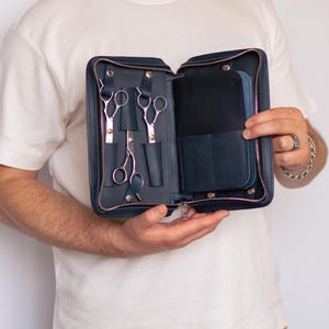 Peut inclure: Un étui zippé en cuir bleu foncé doté de fentes et de sangles internes conçues pour maintenir trois paires de ciseaux de toilettage en métal argenté. L'intérieur comprend une section de poche dédiée pour des outils ou accessoires avec fermeture.