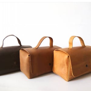 Könnte beinhalten: Drei Ledertaschen in Braun, Tan und Hellbraun mit oberen Griffen. Die Taschen sind alle gleich groß und in Form, und sie sind in einer Reihe von links nach rechts angeordnet.