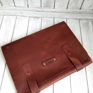 May include: A brown leather laptop sleeve with a personalized name, "Anne", embossed on the front. The sleeve has two straps with brass buckles.