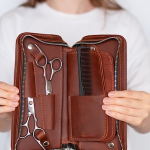 Peut inclure: Étui à outils de coiffeur en cuir marron ouvert avec une fermeture éclair noire. L'étui contient une paire de ciseaux, un peigne et d'autres outils de coiffeur. L'étui est ouvert, révélant les outils à l'intérieur.