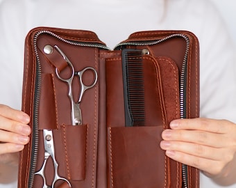 Puede incluir: Estuche de herramientas de peluquería de cuero marrón abierto con cremallera negra. El estuche contiene unas tijeras, un peine y otras herramientas de peluquería. El estuche está abierto, revelando las herramientas en su interior.