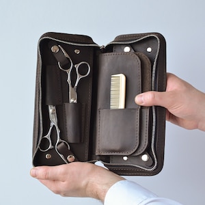 May include: A brown leather zippered pouch with two pairs of scissors and a comb inside. The pouch is open and held by a person's hands.