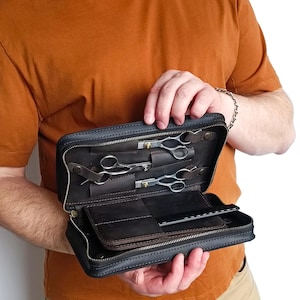 Op de afbeelding: Een donkerbruine lederen etui met ritssluiting met twee paar zilverkleurige metalen kappersscharen en een zwarte kam. De binnenzijde is voorzien van speciale lederen vakjes voor de scharen en een plat vak voor het opbergen van gereedschap.