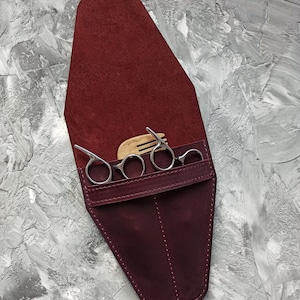 Op de afbeelding: Een bordeauxrode leren etui met een klep sluiting, met een set zilveren kappers scharen en een houten kam.