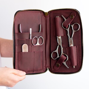 May include: A burgundy leather zippered case with multiple compartments for holding hairdressing tools. The case is open and shows a pair of shears, a comb, and other tools.