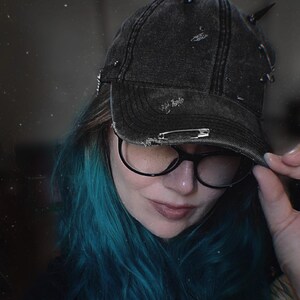 May include: Black denim baseball cap with distressed details and metal spikes. The cap features a safety pin detail and is worn with black-framed glasses and teal hair. A stylish accessory for a unique look.