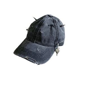 May include: A distressed, dark gray baseball cap with metal spikes and safety pins. The cap features a small, silver charm with a heart and dagger design. The fabric has a worn appearance with small tears.