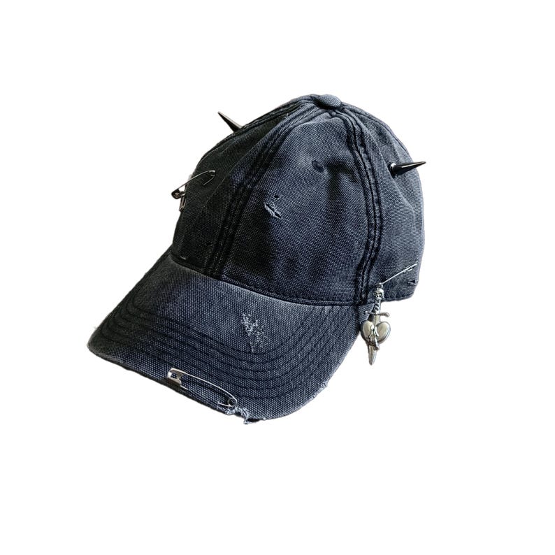 May include: A distressed, dark gray baseball cap with metal spikes and safety pins. The cap features a small, silver charm with a heart and dagger design. The fabric has a worn appearance with small tears.