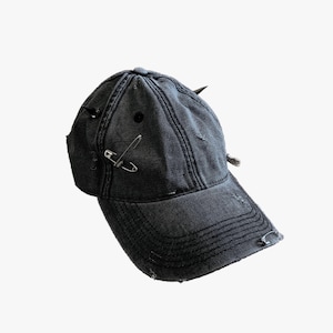 May include: A distressed black baseball cap with a curved brim. The cap features a metal safety pin and small metal spikes. The fabric has a worn appearance with distressed edges and visible stitching.