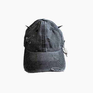 May include: A distressed black baseball cap with two black spikes on top and several safety pins and a charm attached. The cap has a worn, faded appearance with visible distressing.