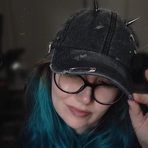 May include: A distressed black denim baseball cap with silver safety pins and metal spikes. The cap has a curved brim and a worn appearance, suggesting a vintage or punk rock aesthetic. The hat is a stylish accessory.