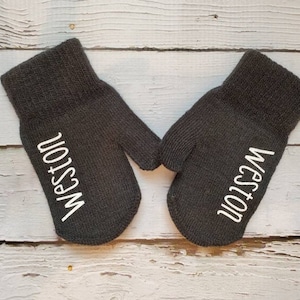 May include: A pair of dark gray knitted mittens with the name "Weston" printed in white on each mitten. The mittens are displayed on a light wood-grain surface.
