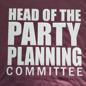 Op de afbeelding: Een bordeauxrood t-shirt met de witte tekst "HEAD OF THE PARTY PLANNING COMMITTEE" op de voorkant. De tekst is in een vetgedrukte, schreefloze lettertype. Het shirt heeft een licht getextureerde uitstraling.